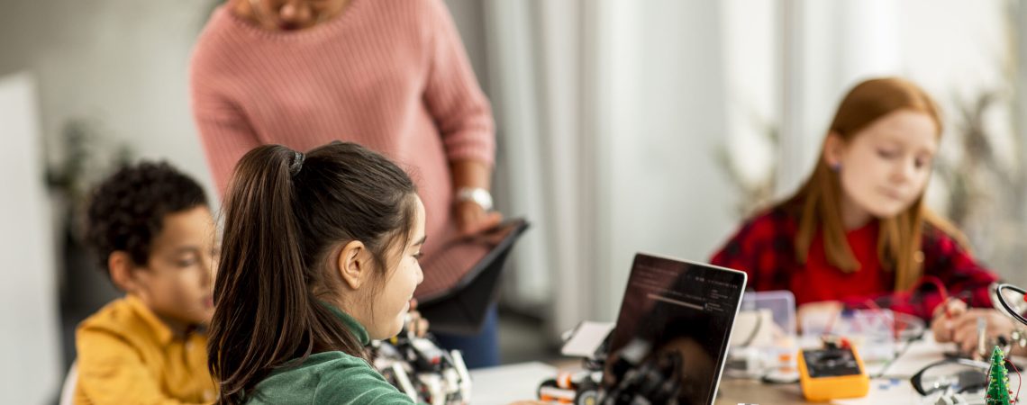 Group of happy kids with their African American female science teacher with laptop programming electric toys and robots at robotics classroom